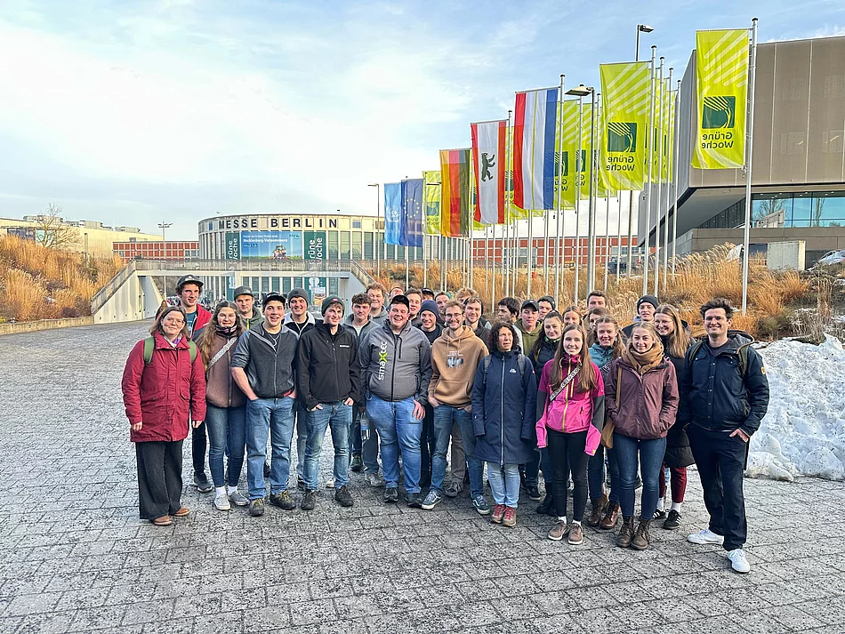 Eine Gruppe von Menschen steht vor einem Großen Gebäude mit der Aufschrift "Messe Berlin". Zu sehen sind auch diverse Fahnen, u.a. Deutschlandfahne, Europafahne sowie einige grüne Fahnen mit der Aufschrift "Grüne Woche".