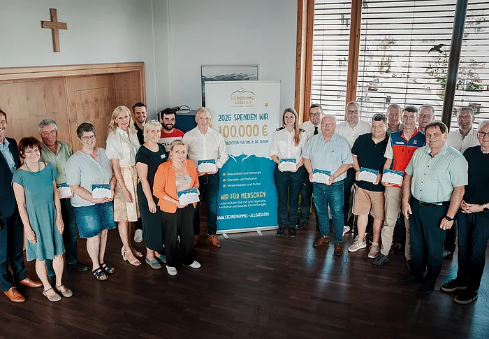 Männer und Frauen stehen in einem Raum für ein Gruppenbild zusammen. Einige von Ihnen halten ein blau/weißes Papier in den Händen und präsentieren es in die Kamera. In der Mitte steht ein Aufsteller.