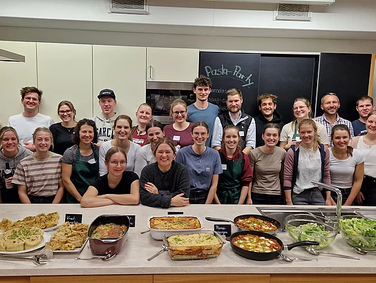 Junge Damen und Herren posieren für ein Gruppenbild in einer Küche. Im Vordergrund auf der Anrichte stehen diverse gekochte Gerichte, angeordnet zu einem kleinen Buffet. Dazwischen stehen auch kleine Täfelchen. Vermutlich sind darauf die Namen der Gerichte vermerkt.