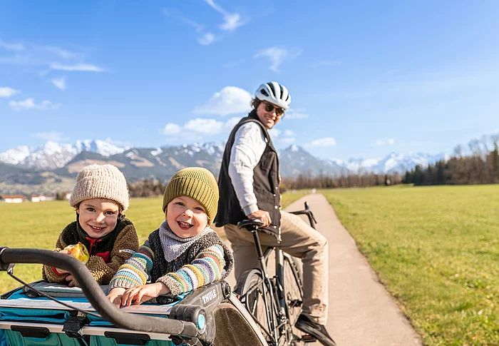 Das Bild zeigt einen Mann auf einem Fahrrad. Er steht und hält das Fahrrad fest, dabei schaut er nach hinten. Am Fahrrad angehängt ist ein Kinderfahrradanhänger. Darin stehen 2 Kinder und lächeln in die Kamera. Die Familie befindet sich auf einem Weg durch Felder. Im Hintergrund sind die Berge zu sehen.