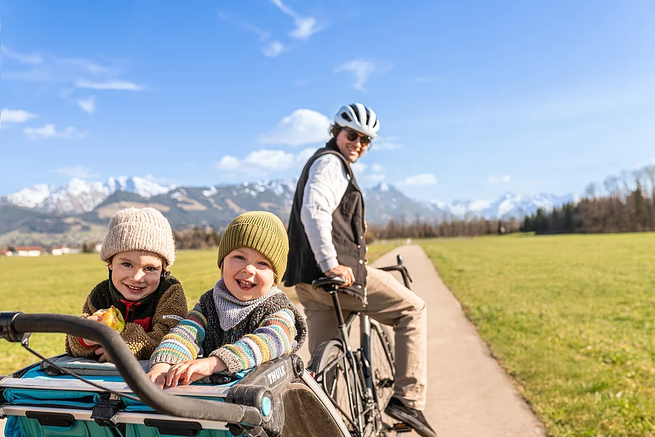 Das Bild zeigt einen Mann auf einem Fahrrad. Er steht und hält das Fahrrad fest, dabei schaut er nach hinten. Am Fahrrad angehängt ist ein Kinderfahrradanhänger. Darin stehen 2 Kinder und lächeln in die Kamera. Die Familie befindet sich auf einem Weg durch Felder. Im Hintergrund sind die Berge zu sehen.