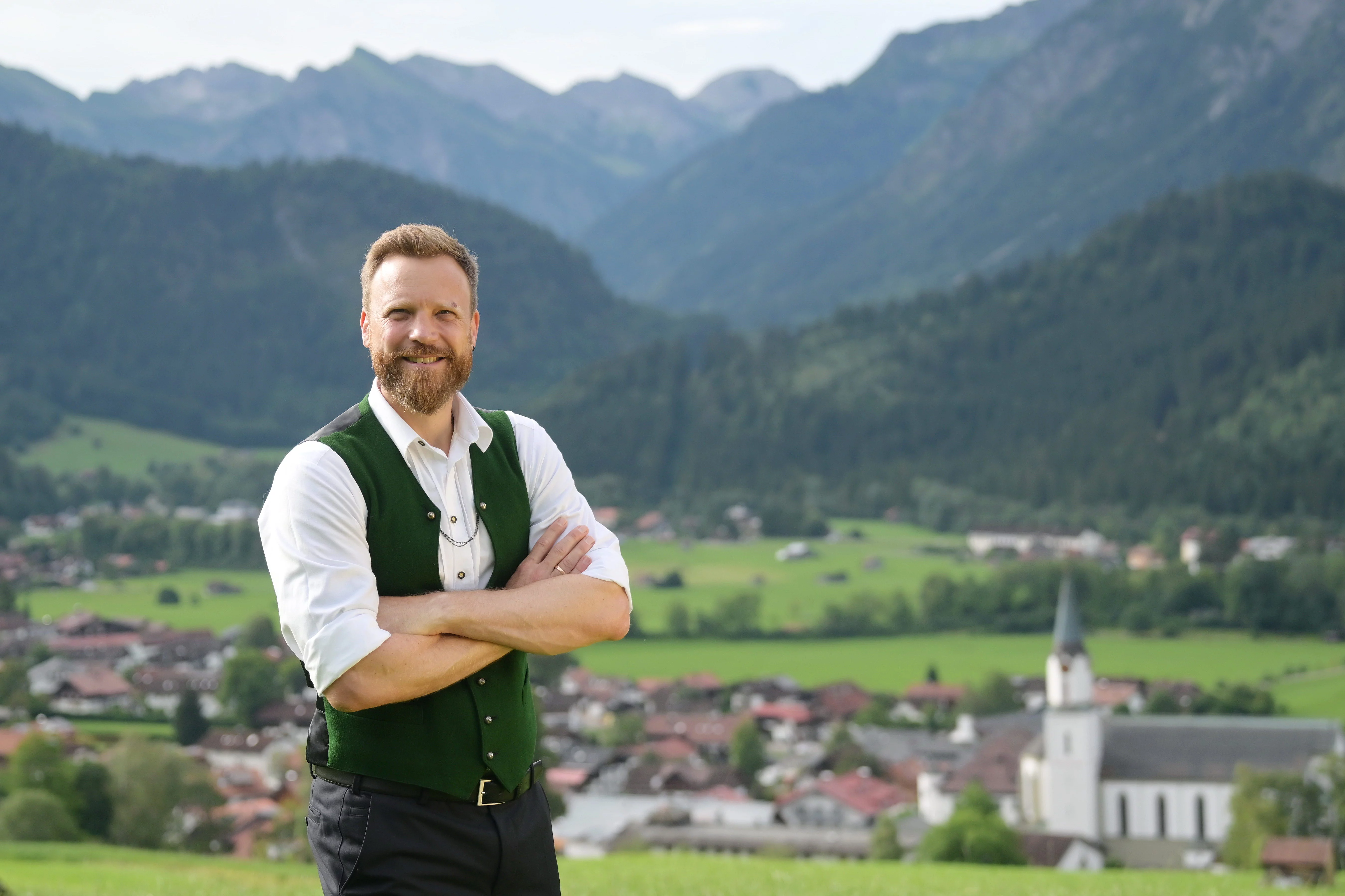 Im Vordergrund steht ein Mann mit verschränkten Händen. Er hat ein weißes Hemd mit grüner Weste und eine schwarze Hose an und lächelt in die Kamera. Im Hintergrund ist verschwommen ein Ort von oberhalb zu sehen. Bei dem Mann handelt es sich um 1. Bürgermeister Tobias Keuschnig.