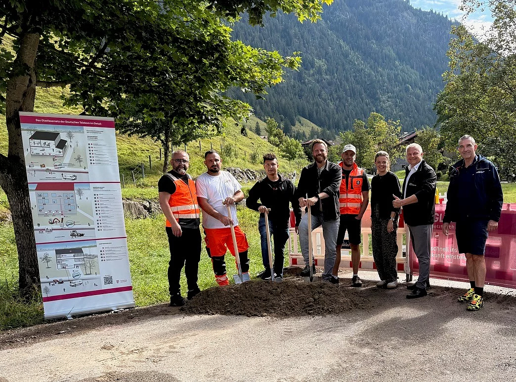 Zu sehen ist eine Menschengruppe auf einem Kiesparkplatz vor einer Baustelle beim Spatenstich