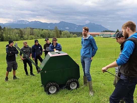 Auf einer grünen Wiese stehen einige junge Männer um ein grünes Gerät mit Rädern herum. Augenscheinlich erklärt ein Dozent die Funktionsweise.